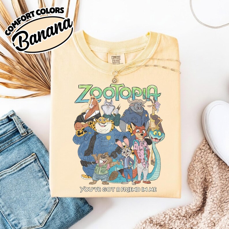 May include: Banana-colored Zootopia t-shirt featuring a group of animated animal characters. The shirt has the text "Zootopia" in green and blue, with the phrase "You've got a friend in me" below. The shirt is part of the Comfort Colors collection.
