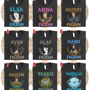 Puede incluir: Nueve camisetas negras con personajes de "Frozen". Cada camiseta muestra el nombre de un personaje, "EST. 2013" y "FROZEN". Los personajes incluyen a Elsa, Anna, Kristoff, Sven, Olaf, Hans, Oaken, Pabbie y Marshmallow.