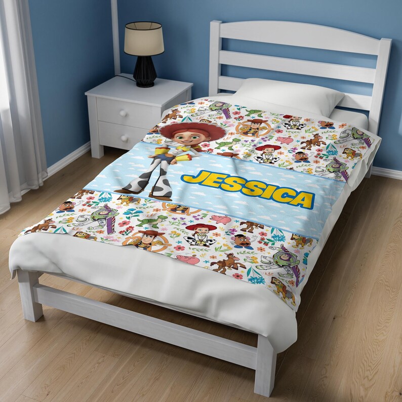 May include: A white bed blanket featuring a repeating pattern of Toy Story characters including Woody, Buzz Lightyear, and Jessie. A central light blue horizontal band displays the name JESSICA in bold yellow letters across the middle.