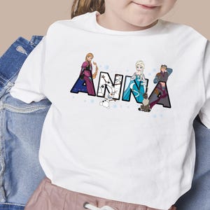 Puede incluir: Camiseta blanca con el nombre "ANNA" en letras grandes y decoradas. Las letras presentan personajes de la pel&iacute;cula Frozen, incluyendo a Anna, Elsa y Kristoff. El dise&ntilde;o est&aacute; sobre un fondo de peque&ntilde;as estrellas azules.