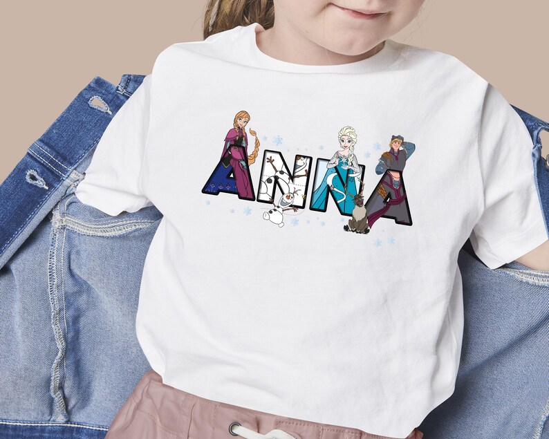 Puede incluir: Camiseta blanca con el nombre "ANNA" en letras grandes y decoradas. Las letras presentan personajes de la pel&iacute;cula Frozen, incluyendo a Anna, Elsa y Kristoff. El dise&ntilde;o est&aacute; sobre un fondo de peque&ntilde;as estrellas azules.