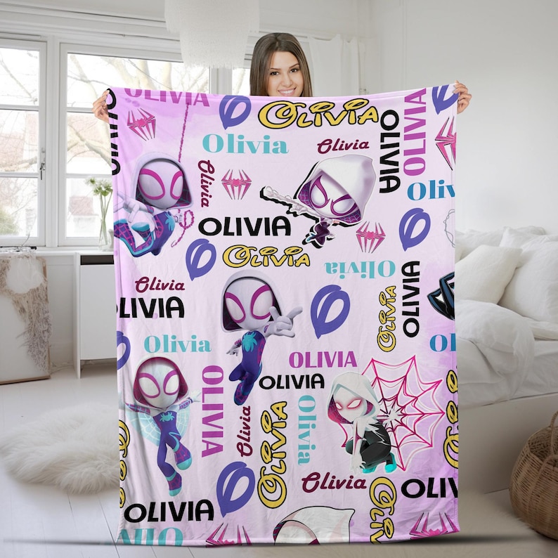 May include: A light pink fleece blanket featuring a repeating pattern of Ghost-Spider characters in various poses, pink spider web graphics, and the name Olivia printed in multiple fonts, colors, and orientations across the entire fabric surface.