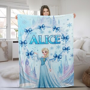 May include: A light blue fleece blanket featuring a printed illustration of a blonde character in a long blue gown, a castle silhouette, snowflakes, and blue bows. The name ALICE is printed in large blue letters in the center of the design.