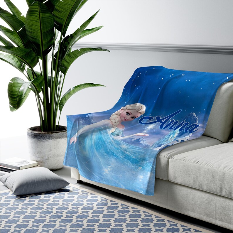 May include: A soft blue throw blanket featuring a graphic print of a blonde character in a light blue dress with the name Anna written in a cursive script. The design includes a starry night sky background with swirling ice and snow elements.