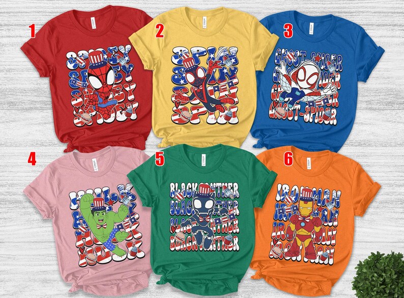 K&ouml;nnte beinhalten: Sechs kurz&auml;rmelige T-Shirts in verschiedenen Farben, jedes mit einem Superhelden-Motiv mit patriotischen Themen. Die Shirts zeigen Charaktere wie Spider-Man, Ghost-Spider, Hulk, Black Panther und Iron Man.