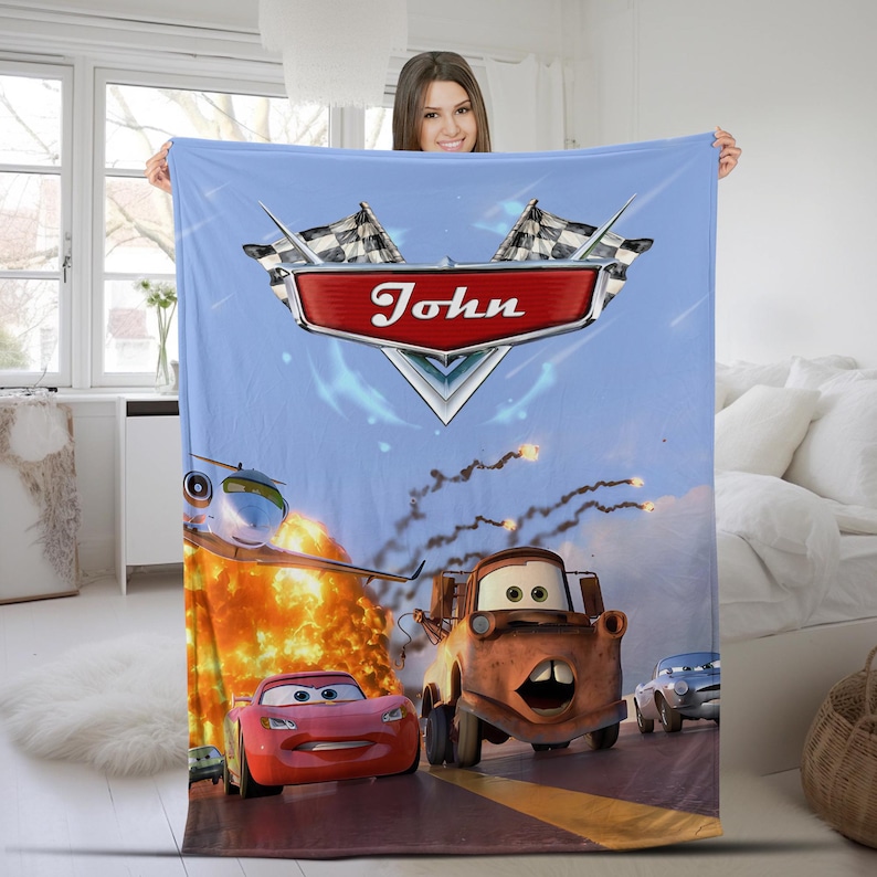 May include: A light blue fleece blanket featuring a graphic print of characters from the movie Cars, including a red race car and a brown tow truck. The design includes a red emblem with the name John and two checkered flags at the top.