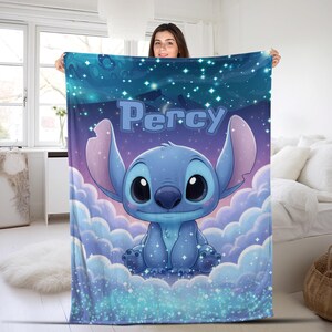 Puede incluir: Una manta rectangular de forro polar con un personaje alien&iacute;gena azul sentado entre nubes azul claro sobre un cielo nocturno estrellado azul oscuro. El nombre Percy est&aacute; impreso en letras grandes de color azul claro sobre el personaje.