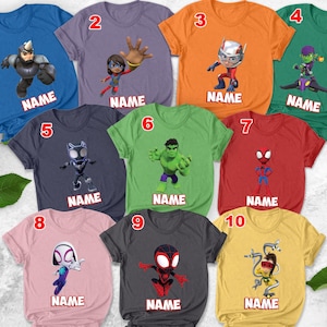 May include: A collection of ten colorful t-shirts featuring various superhero characters. Each shirt has a different design, including characters like Spider-Man and Hulk, with the word "NAME" printed below each character. The shirts are in various colors, including blue, orange, and green.