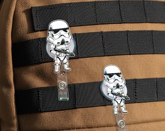 May include: Two white Stormtrooper badge reels with black and silver details are attached to a brown backpack. Each reel features a Stormtrooper figure holding a blaster. The reels have clear plastic straps and circular attachments.