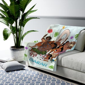 K&ouml;nnte beinhalten: Eine hellblaue Fleecedecke mit einem farbenfrohen Grafikdruck von animierten Charakteren aus dem Film Vaiana, darunter Vaiana, Maui, Pua und Heihei, verziert mit tropischen Blumen und Bl&auml;ttern, mit dem in Blau gedruckten Namen Sarah.