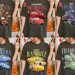 May include: Dark grey t-shirts featuring characters from the animated film Cars. Designs include Tow Mater, Lightning McQueen, Doc Hudson, Sally Carrera, Cruz Ramirez, and Fillmore, with lightning bolt graphics.