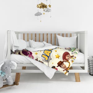 May include: A white baby blanket featuring a yellow striped pattern, gold stars, and characters from Toy Story including Woody, Jessie, and Lotso. The name JACK is printed in large blue letters with a yellow outline across the center of the blanket.