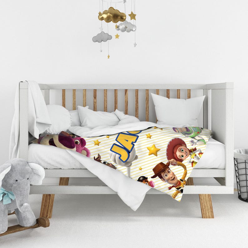 May include: A white baby blanket featuring a yellow striped pattern, gold stars, and characters from Toy Story including Woody, Jessie, and Lotso. The name JACK is printed in large blue letters with a yellow outline across the center of the blanket.