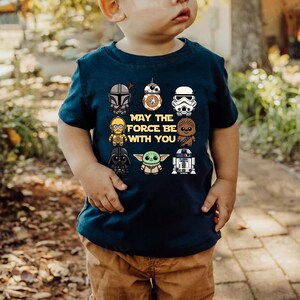 May include: Navy blue t-shirt featuring various Star Wars characters and the phrase "MAY THE FORCE BE WITH YOU." The shirt showcases characters like Darth Vader, Yoda, and R2-D2 in a cartoon style. The shirt is being worn by a child.