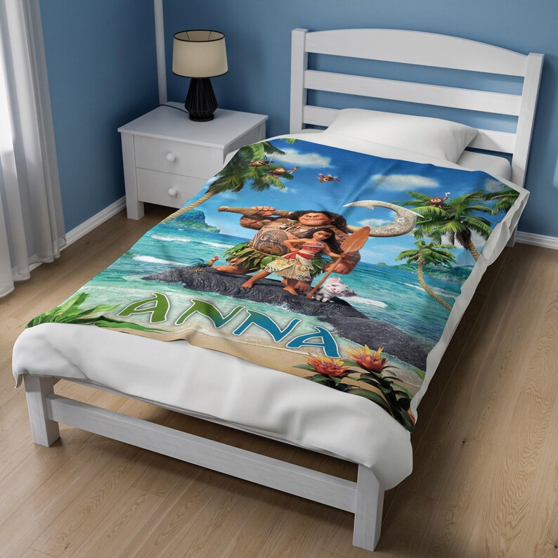 Op de afbeelding: Een wit bedframe met matras en een deken met een geprinte sc&egrave;ne van Moana en Maui op een tropisch strand. De deken toont de naam ANNA in grote blauwe letters, palmbomen, oceaangolven en kleurrijke bloemen.