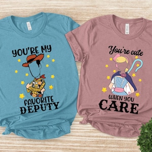 May include: Two t-shirts, one light blue and one mauve, with cartoon graphics. The light blue shirt says "You're my favorite deputy" with a Woody character. The mauve shirt says "You're cute when you care" with a Bo Peep character.
