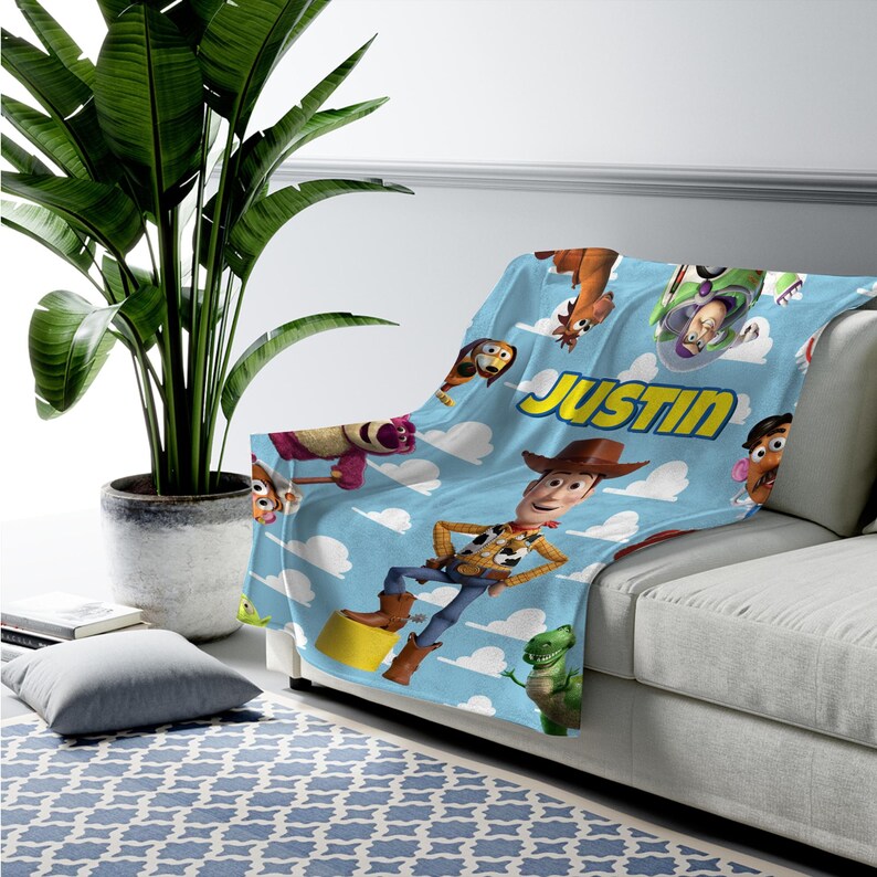 K&ouml;nnte beinhalten: Eine hellblaue Fleecedecke mit einem wiederkehrenden Muster aus Toy Story-Figuren wie Woody, Buzz Lightyear, Rex und Slinky Dog vor einem Himmelshintergrund mit wei&szlig;en Wolken. Der Name JUSTIN ist in gelber Schrift auf die Decke gedruckt.