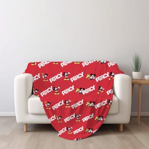 Puede incluir: Una manta roja con un patr&oacute;n repetitivo de Mickey Mouse en varias poses junto al nombre PERCY impreso en letras may&uacute;sculas blancas y negritas. La manta est&aacute; colocada sobre un sof&aacute; blanco, mostrando un dise&ntilde;o gr&aacute;fico vibrante y consistente.