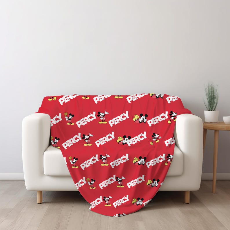Puede incluir: Una manta roja con un patr&oacute;n repetitivo de Mickey Mouse en varias poses junto al nombre PERCY impreso en letras may&uacute;sculas blancas y negritas. La manta est&aacute; colocada sobre un sof&aacute; blanco, mostrando un dise&ntilde;o gr&aacute;fico vibrante y consistente.