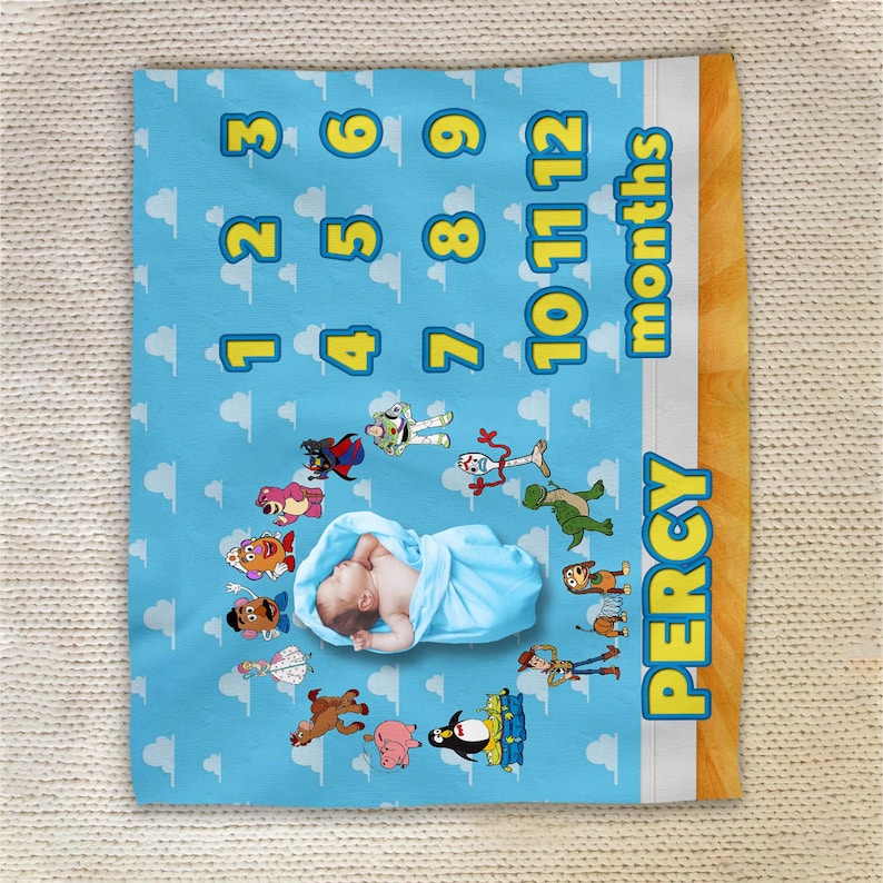 May include: A light blue milestone blanket featuring a repeating cloud pattern, the numbers 1 through 12, the word months, and the name PERCY in yellow. The blanket displays various cartoon characters arranged in a circle around a central infant.