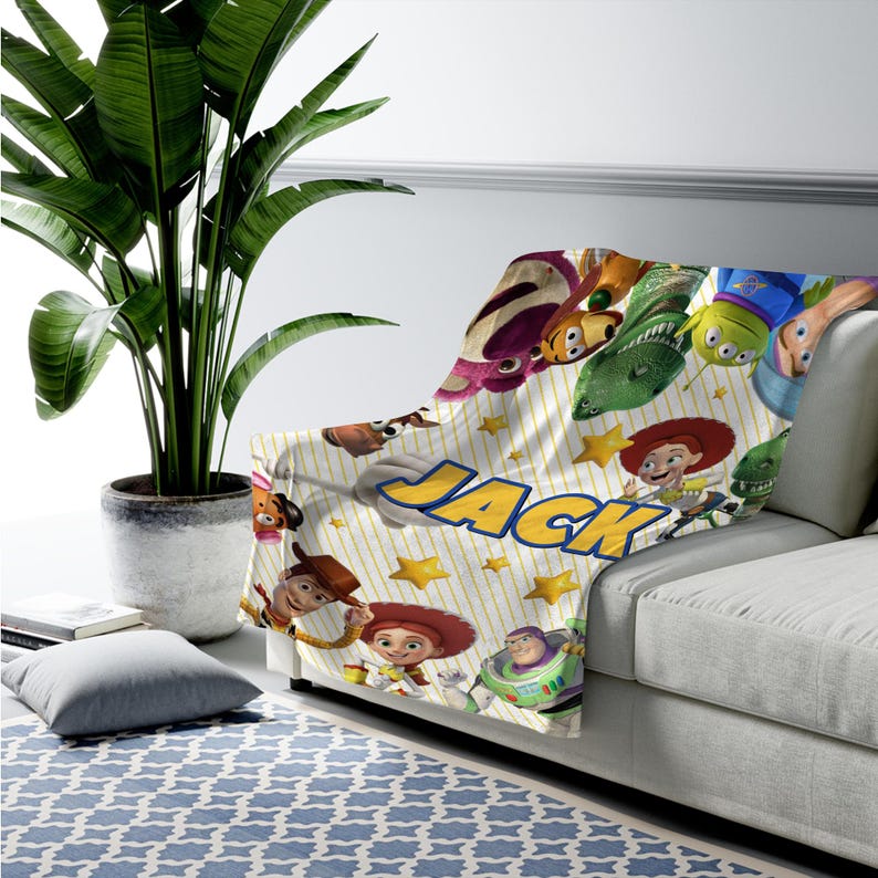 May include: A white fleece blanket featuring a repeating pattern of Toy Story characters including Woody, Buzz Lightyear, Jessie, and Rex, accented with yellow stars and thin vertical stripes, with the name JACK printed in large blue letters.