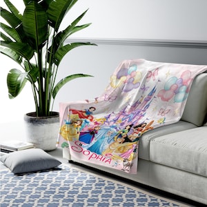 May include: A soft throw blanket featuring a light pink background with a printed illustration of various animated princesses, a large castle, colourful balloons, and butterflies. The name Sophia is printed in large pink script at the bottom.