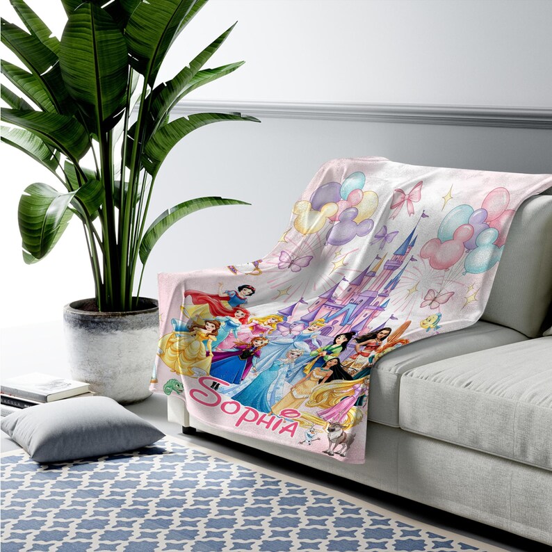 May include: A soft throw blanket featuring a light pink background with a printed illustration of various animated princesses, a large castle, colourful balloons, and butterflies. The name Sophia is printed in large pink script at the bottom.