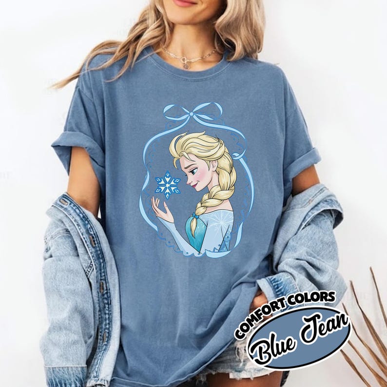 May include: Blue Jean Comfort Colors t-shirt featuring a cartoon illustration of a blonde woman with a snowflake. The woman is wearing a blue dress and has a long braid. The shirt has rolled-up sleeves and the words "COMFORT COLORS" and "Blue Jean" are printed on the bottom right.