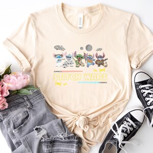 May include: A beige t-shirt featuring a graphic of several cartoon characters with lightsabers and the text "STITCH WARS". The shirt is paired with gray jeans and black canvas sneakers.
