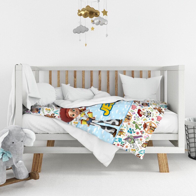 May include: A white crib featuring a blanket with a Toy Story theme, displaying characters like Jessie, Woody, and Buzz Lightyear alongside the name JESS. The blanket has a light blue section with clouds and a white section with a colorful character pattern.