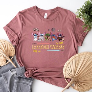May include: Dusty rose t-shirt featuring a graphic of several cartoon characters in a Star Wars theme, with the text "Stitch Wars" in yellow. The design includes lightsabers and spaceships. The shirt is folded.