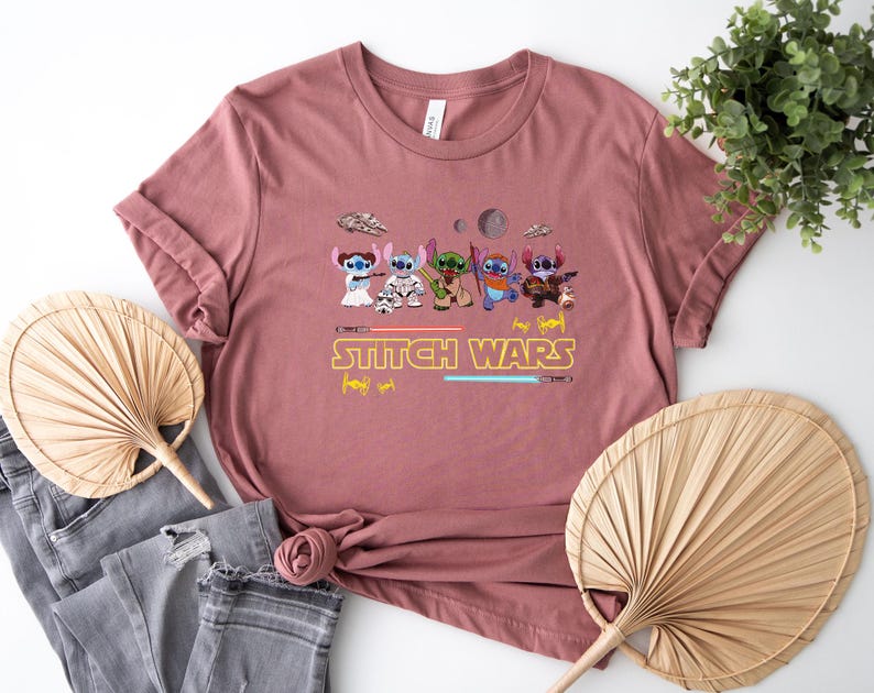 May include: Dusty rose t-shirt featuring a graphic of several cartoon characters in a Star Wars theme, with the text "Stitch Wars" in yellow. The design includes lightsabers and spaceships. The shirt is folded.
