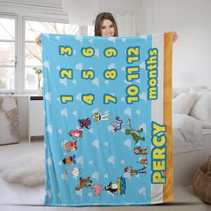 May include: A light blue milestone blanket featuring a repeating white cloud pattern, yellow numbers 1 through 12, and the word months. The blanket displays various cartoon characters arranged in a circle and the name PERCY in yellow letters.