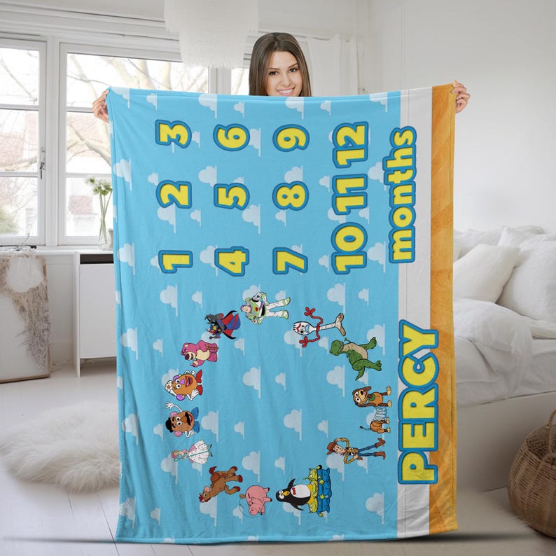 May include: A light blue milestone blanket featuring a repeating white cloud pattern, yellow numbers 1 through 12, and the word months. The blanket displays various cartoon characters arranged in a circle and the name PERCY in yellow letters.