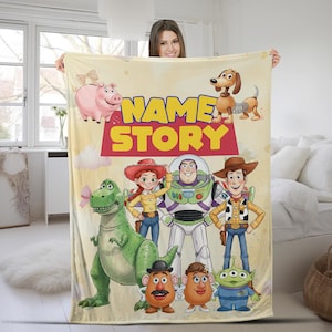 May include: A cream-colored fleece blanket featuring the Toy Story characters Hamm, Slinky Dog, Rex, Jessie, Buzz Lightyear, Woody, Mr. Potato Head, Mrs. Potato Head, and an Alien. The text "NAME STORY" is printed in large yellow and red letters.