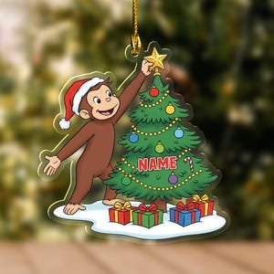 May include: A clear acrylic Christmas ornament featuring a cartoon monkey wearing a Santa hat, reaching for a star atop a Christmas tree. The tree is decorated with ornaments and lights, with the word "NAME" on it. Presents are at the base.