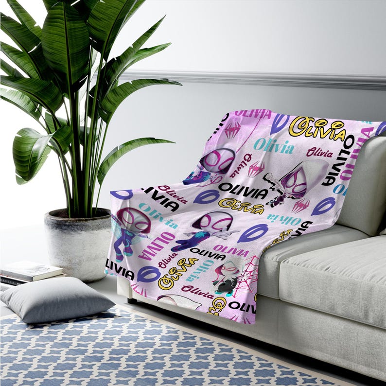 May include: A soft throw blanket featuring a repeating pattern of the name Olivia in various fonts and colors, interspersed with illustrations of a masked superhero character in a white, purple, and black suit, along with spider web motifs.