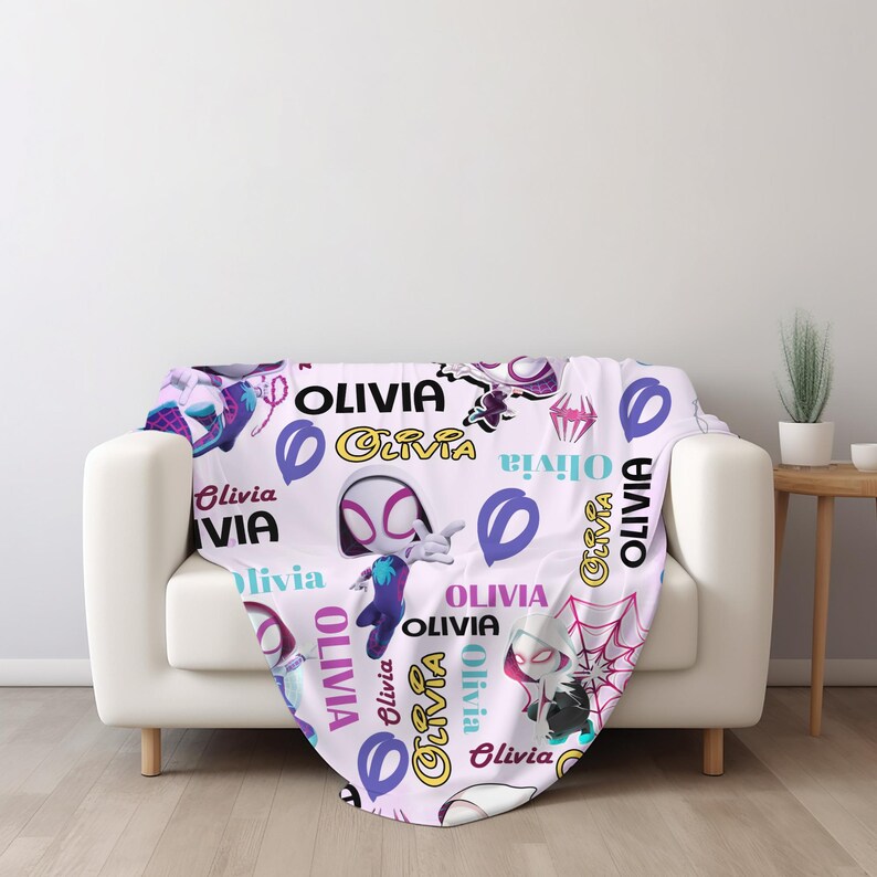 May include: A light purple throw blanket featuring a repeating pattern of the name Olivia in various fonts and colors, interspersed with cartoon illustrations of a masked superhero character in a white, black, and purple suit and spider web motifs.