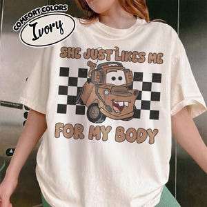 May include: An ivory t-shirt featuring a graphic of a brown tow truck character with a headset. The design includes a black and white checkered background and the text "SHE JUST LIKES ME FOR MY BODY" in brown letters. A label reads "COMFORT COLORS IVORY".