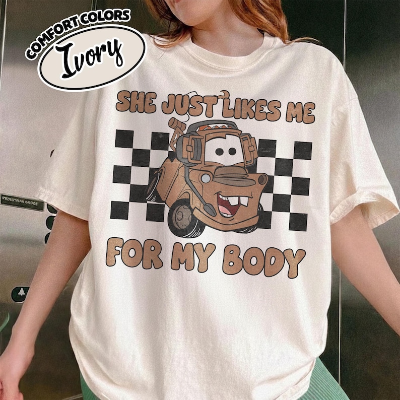 May include: An ivory t-shirt featuring a graphic of a brown tow truck character with a headset. The design includes a black and white checkered background and the text "SHE JUST LIKES ME FOR MY BODY" in brown letters. A label reads "COMFORT COLORS IVORY".