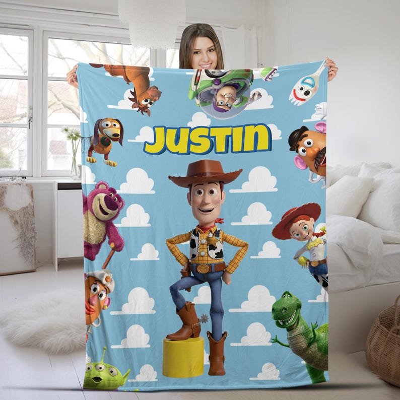 K&ouml;nnte beinhalten: Eine hellblaue Fleecedecke mit einem wiederkehrenden wei&szlig;en Wolkenmuster und verschiedenen Toy Story-Figuren wie Woody, Buzz Lightyear, Jessie, Rex und Slinky Dog. Der Name JUSTIN ist in gro&szlig;en gelben Buchstaben oben in der Mitte aufgedruckt.