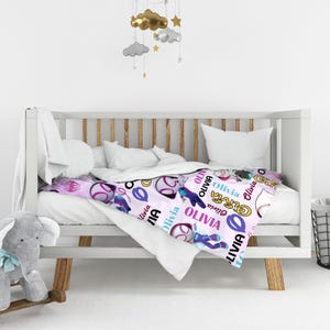 May include: A light pink baby blanket featuring a repeating pattern of cartoon superhero characters, circular icons, and the name Olivia printed in various colors and fonts. The blanket is draped over a white wooden crib with white bedding.