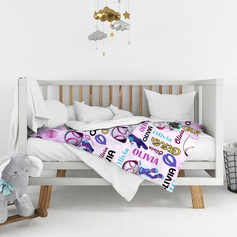 May include: A light pink baby blanket featuring a repeating pattern of cartoon superhero characters, circular icons, and the name Olivia printed in various colors and fonts. The blanket is draped over a white wooden crib with white bedding.