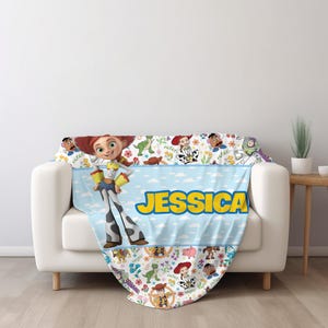 May include: A soft throw blanket featuring a light blue center stripe with the name JESSICA in bold yellow letters. The design includes Toy Story characters like Jessie, Buzz Lightyear, and Rex, surrounded by colorful floral patterns on white.