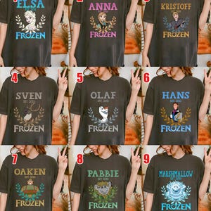 Puede incluir: Nueve camisetas de color gris oscuro con personajes de la pel&iacute;cula "Frozen". Cada camiseta tiene el nombre de un personaje, el texto "EST. 2013" y la palabra "FROZEN" en una fuente decorativa. Los personajes incluyen Elsa, Anna, Kristoff, Sven, Olaf, Hans, Oaken, Pabbie y Marshmallow.