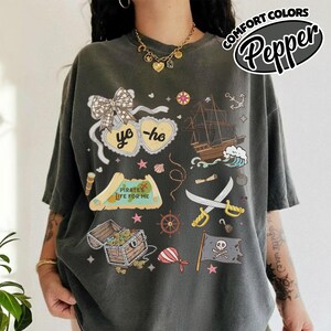 May include: Dark gray t-shirt with a pirate-themed graphic. The design includes a treasure chest, ship, map, crossed swords, and the text "Pirate's Life for Me." The shirt also features the text "yo ho" in a heart shape and the words "Comfort Colors Pepper".