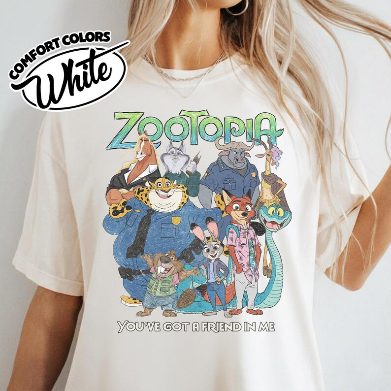 May include: White Comfort Colors t-shirt featuring a colorful graphic of characters from the animated movie Zootopia. The text "Zootopia" is in green, and the phrase "You've Got a Friend in Me" is below the graphic.