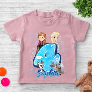 Pode incluir: Camiseta rosa claro infantil com um design de Frozen. Apresenta Anna e Elsa, um grande n&uacute;mero 4 azul e o nome "Sophia" em it&aacute;lico. Olaf e Sven tamb&eacute;m s&atilde;o representados. Ideal para o quarto anivers&aacute;rio.