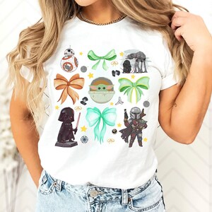 May include: White t-shirt featuring Star Wars characters and decorative bows. The design includes illustrations of BB-8, Darth Vader, The Mandalorian, and Baby Yoda, along with green and brown bows and star accents.