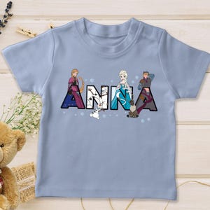 Puede incluir: Una camiseta azul claro con el nombre "ANNA" impreso en letras grandes y coloridas. Cada letra est&aacute; decorada con personajes de la pel&iacute;cula Frozen, incluyendo a Anna, Elsa, Kristoff y Olaf. La camiseta est&aacute; hecha de un material suave.